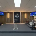 Completed view of the renovated JCS stage, highlighting the central section of custom slat wall panels with integrated LED lighting and pro-AV equipment.