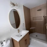 Completed modern bathroom remodel showing a wood vanity, brass mirror, and wood slat shower tiles.