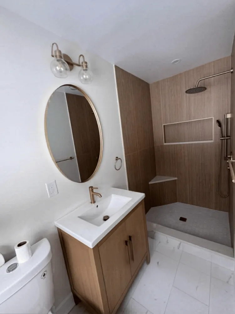 Completed modern bathroom remodel showing a wood vanity, brass mirror, and wood slat shower tiles.